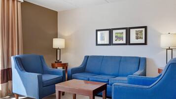 A hotel room with a blue armchair, a blue sofa, a wooden coffee table, and two floor lamps. There are three framed pictures on the wall.
