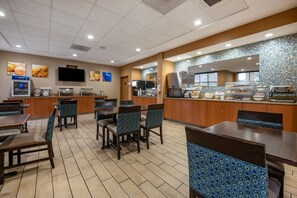 A restaurant with wooden tables and blue patterned chairs, a counter with food display, and a tiled backsplash. - Comfort Inn & Suites Orlando North (Sanford)