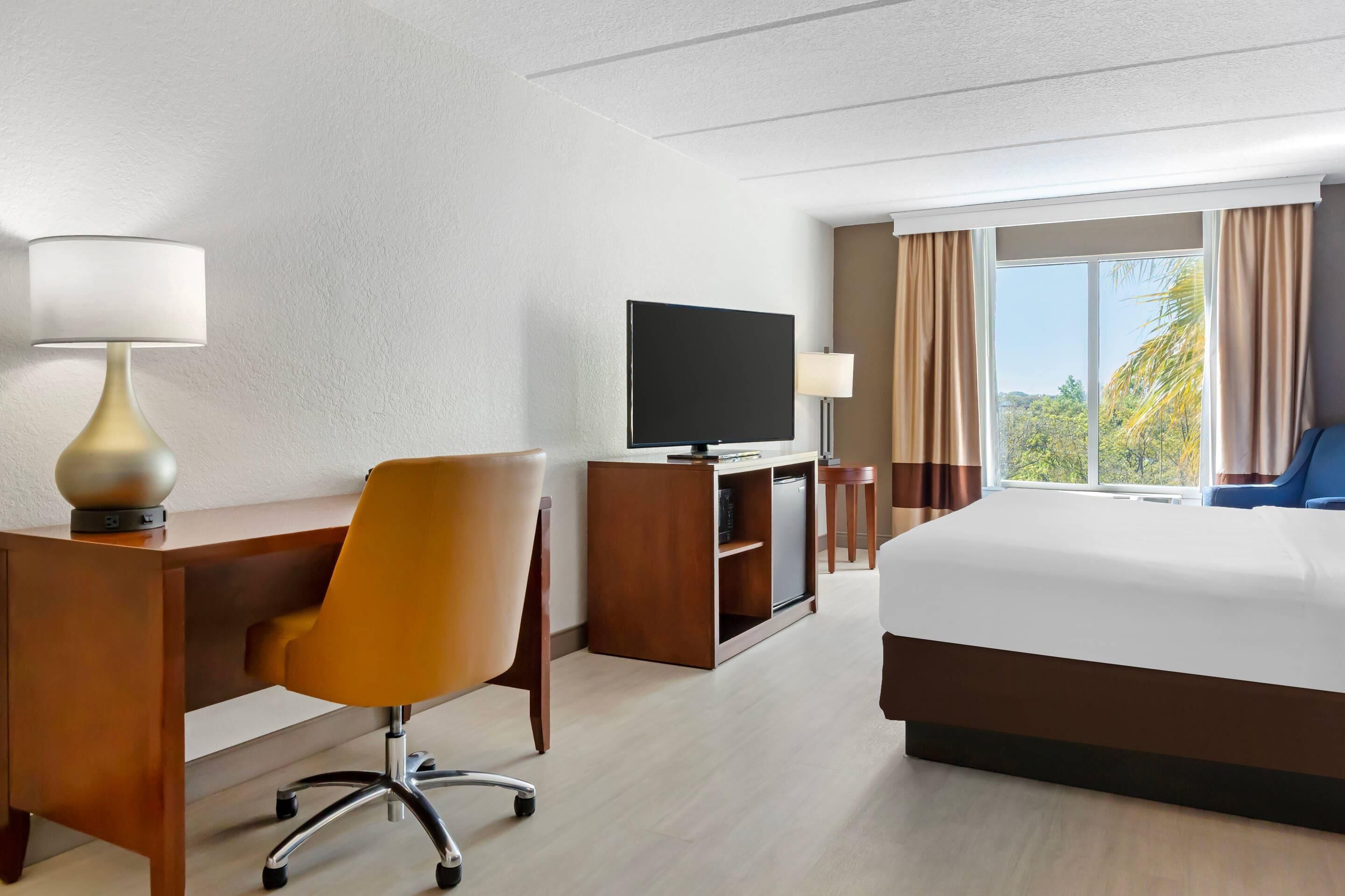 A hotel room with a bed, a desk with a chair, a television, and a window with curtains.