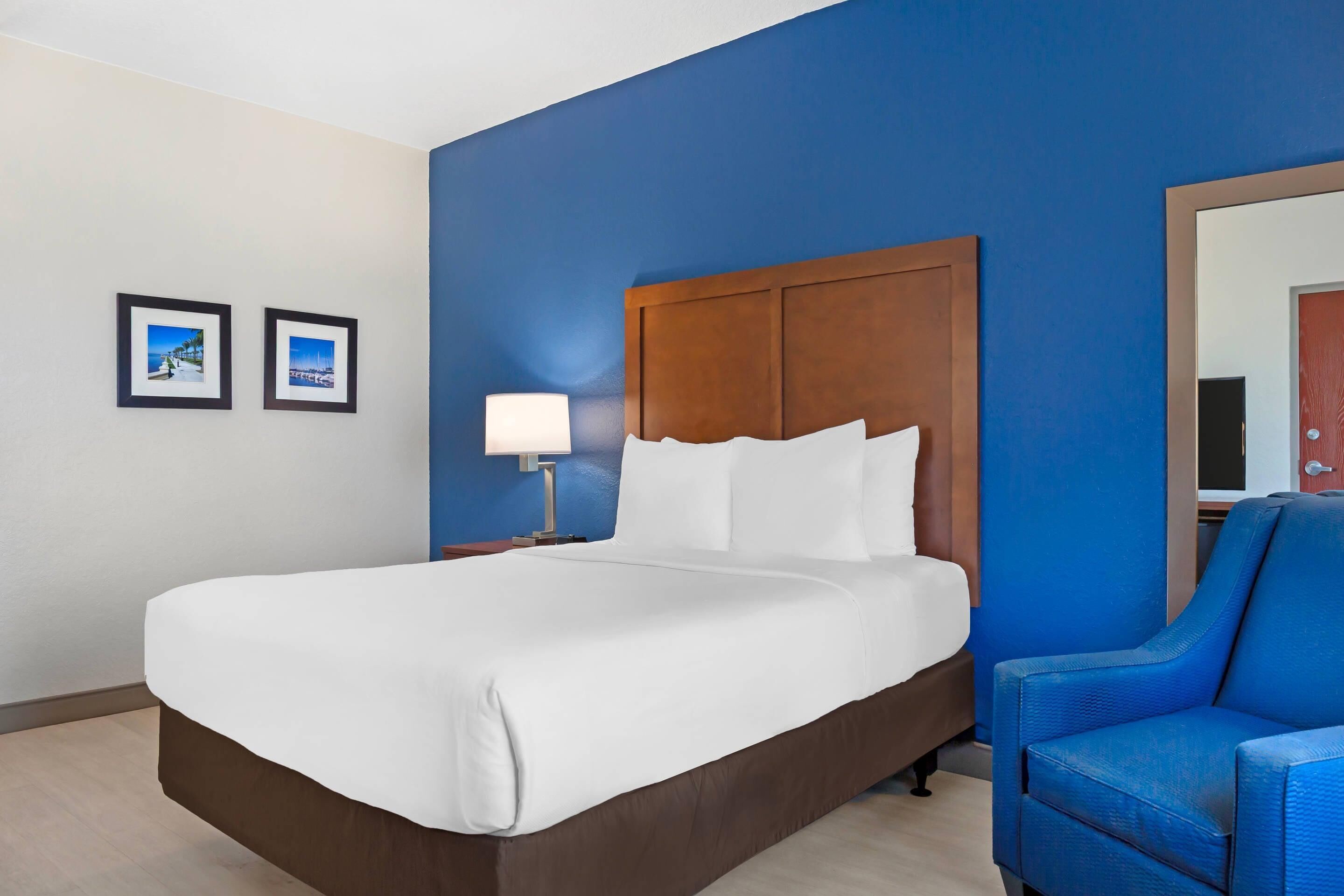 Quarto de hotel com uma cama grande, cabeceira de madeira, parede azul, uma poltrona azul e duas imagens emolduradas na parede.
