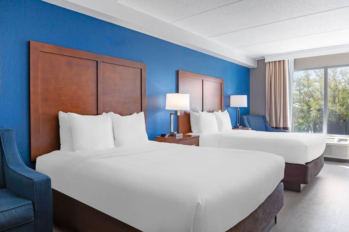 A hotel room with two beds, a blue accent wall, and a large window with a view of trees.
