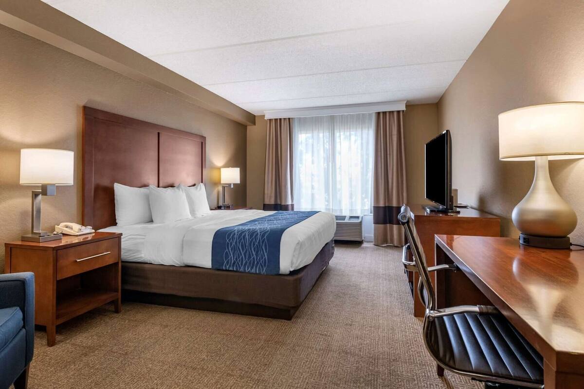 A hotel room with a bed, bedside tables, a desk, a chair, a TV, and a window with curtains.