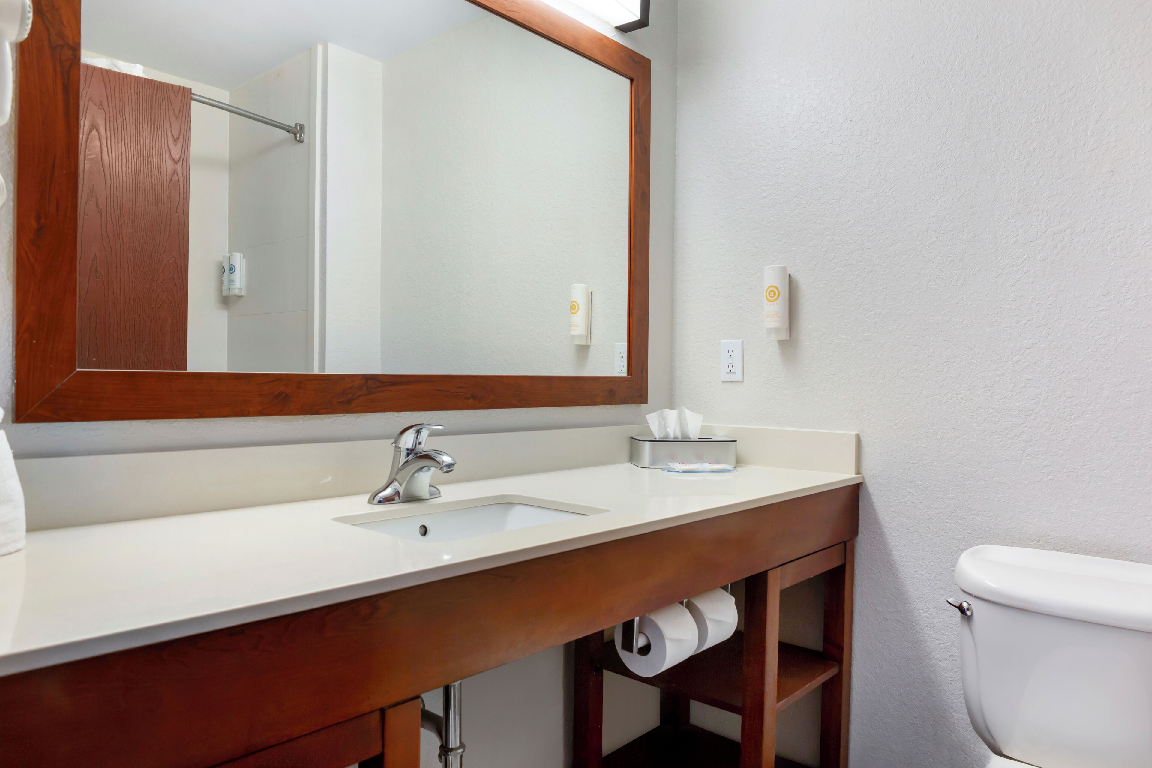 Un baño con tocador de madera, lavamanos, espejo, inodoro y dispensador de toallas.