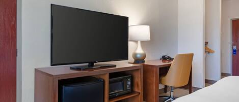 Habitación de hotel con televisión de pantalla plana, lámpara, microondas, refrigerador pequeño y una silla.