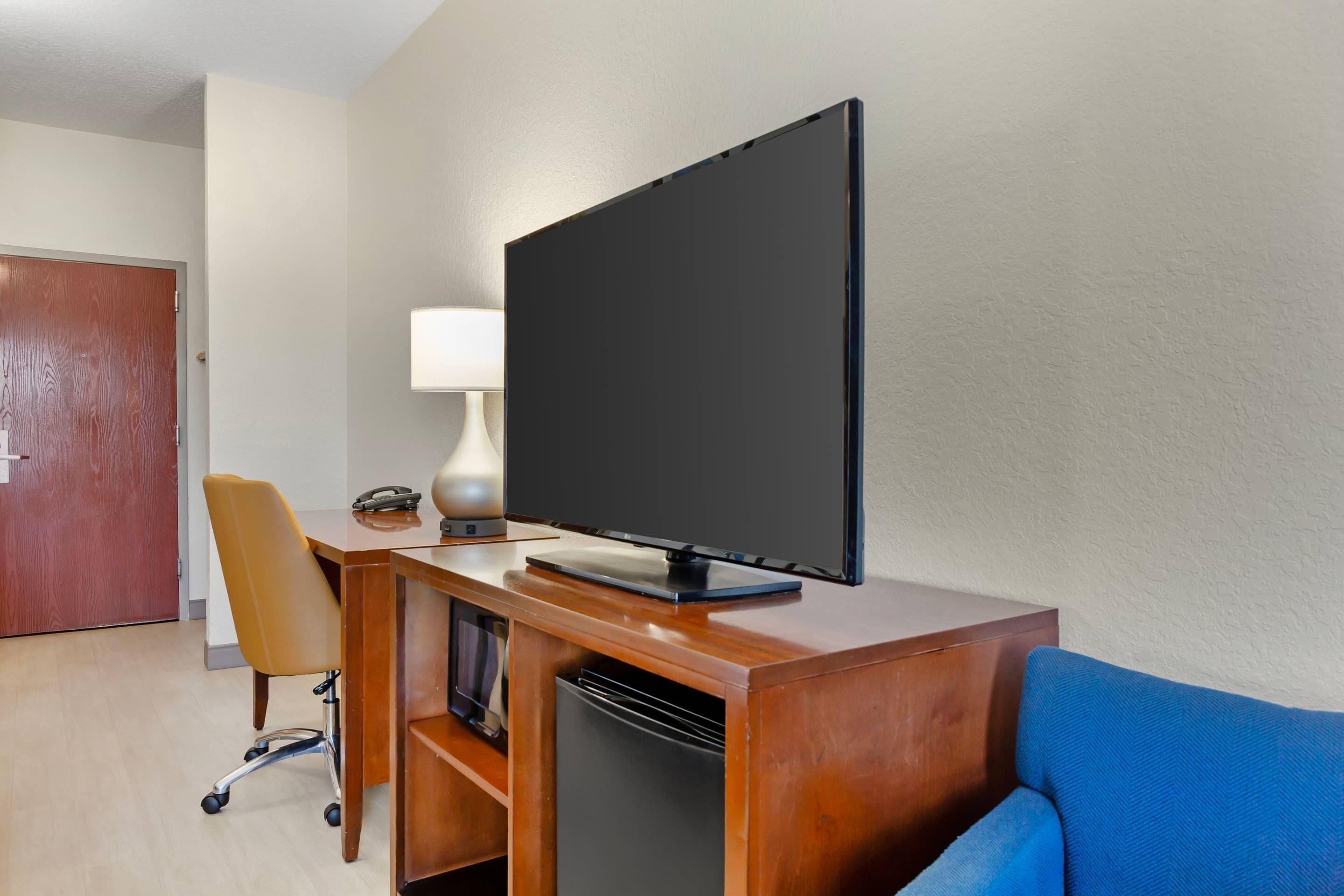 Una habitación de hotel con un escritorio de madera, televisión, una silla, lámpara y un sofá azul.