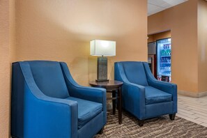 A hotel lobby with two blue armchairs, a small table with a lamp, and a refrigerator stocked with beverages. - Comfort Inn & Suites Orlando North (Sanford)