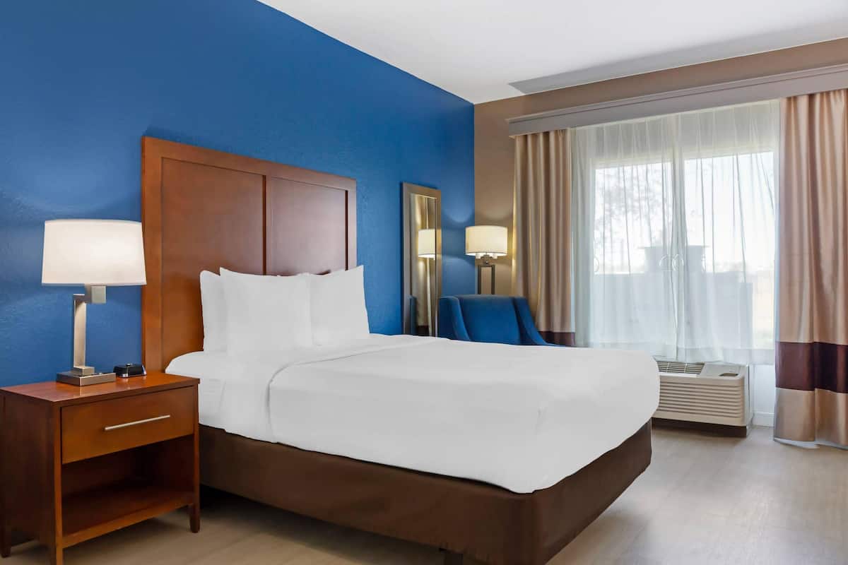 A hotel room with a large bed, a nightstand with a lamp, a mirror, and an air conditioning unit.