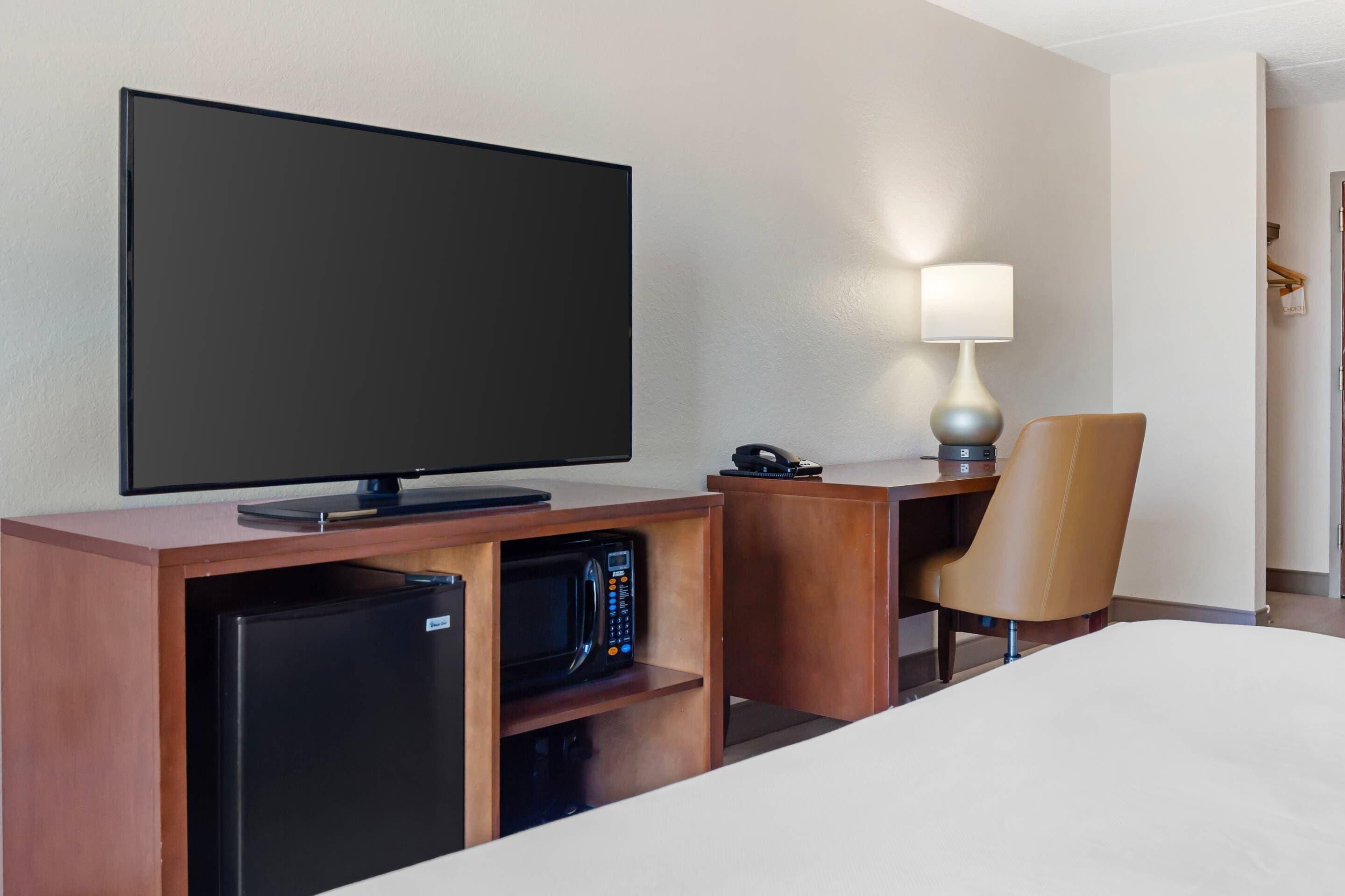 A hotel room with a flat-screen TV, a desk with a lamp, a microwave, a refrigerator, and a bed.