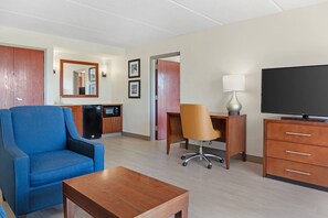 A hotel room with a blue armchair, a wooden desk, a television, and a minibar. - Comfort Inn & Suites Orlando North (Sanford)