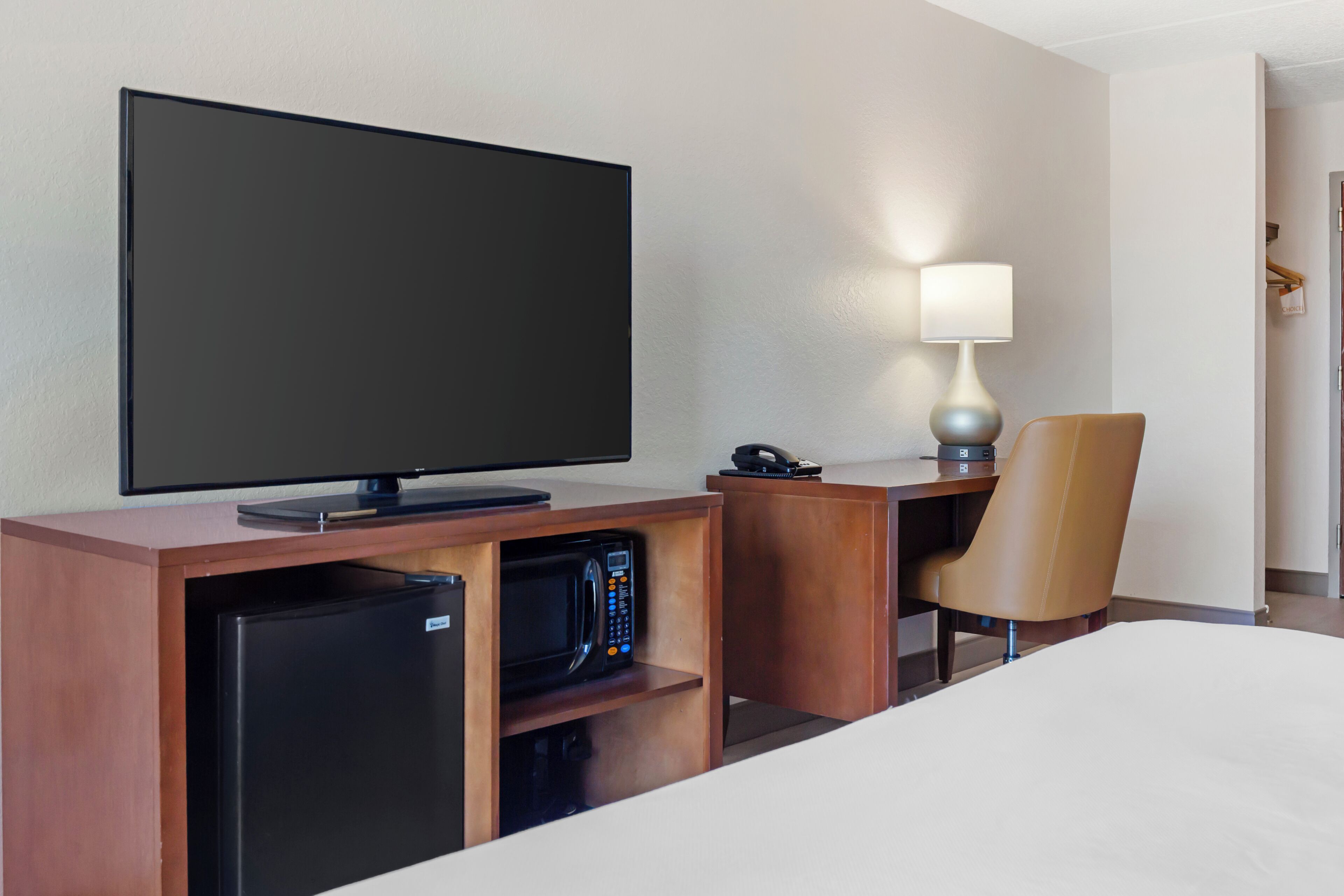 A hotel room with a flat-screen TV, a desk with a lamp, a microwave, and a chair.