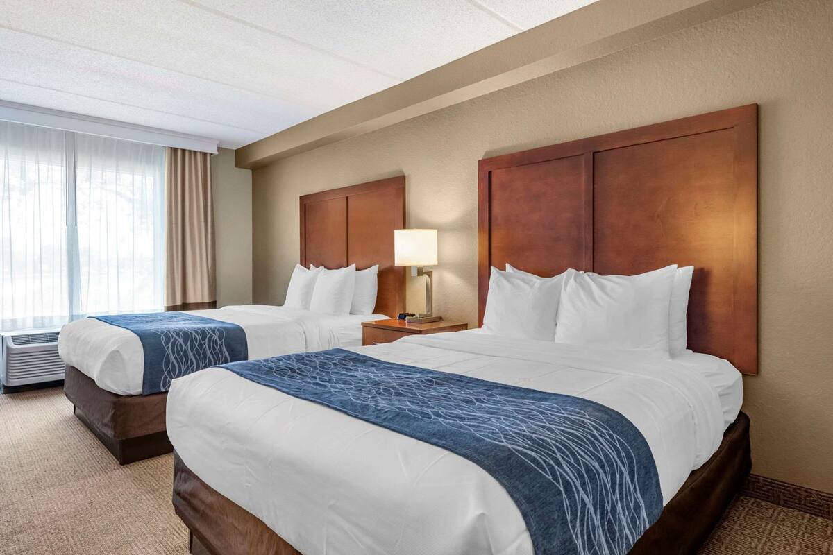 A hotel room with two beds, a nightstand with a lamp, a window with curtains, and a ceiling with a light fixture.