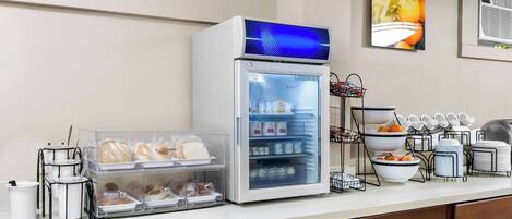 Un espace petit-déjeuner à l’hôtel, équipé d’un réfrigérateur rempli de nourriture, d’une machine à café et de divers articles pour le petit-déjeuner disposés sur le comptoir.