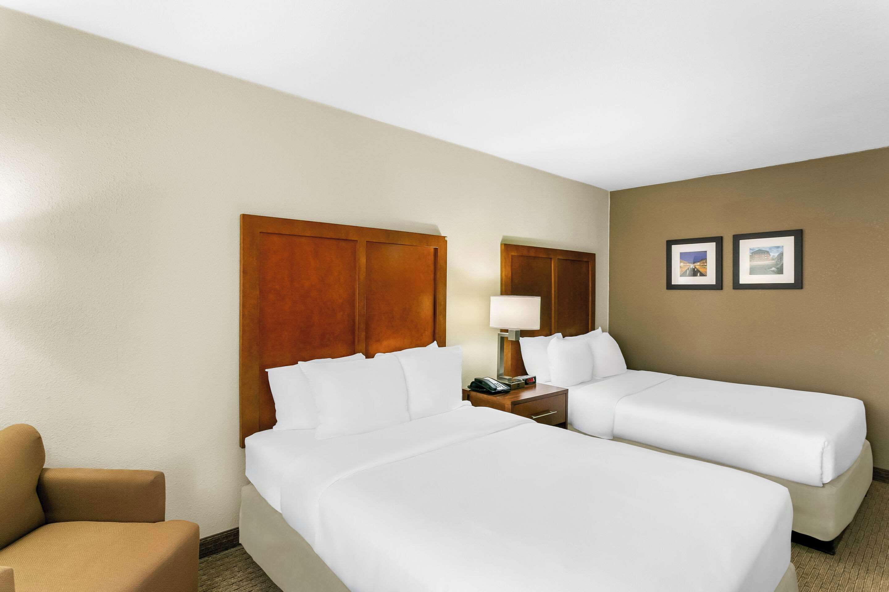 A hotel room with two beds, a wooden headboard, a bedside table with a lamp, and two framed pictures on the wall.