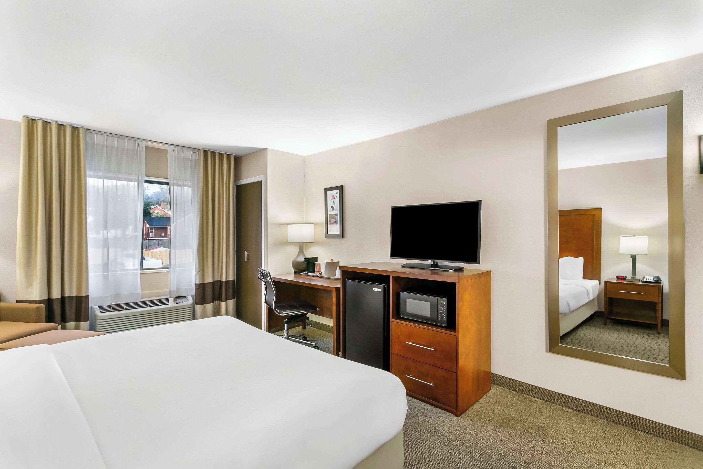 A hotel room with a bed, a desk with a TV, a refrigerator, a lamp, and a large mirror.