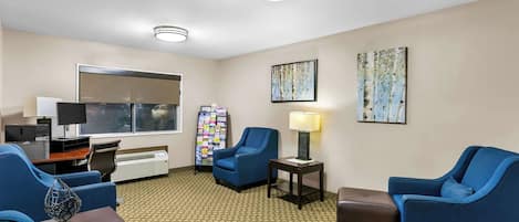 Habitación de hotel con un ventanal grande, un escritorio con computadora, dos sillones azules, una mesita con lámpara y un tablón de anuncios con varios objetos.