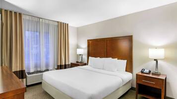 A hotel room with a large bed, a wooden nightstand with a lamp, a heater, and a window with curtains.