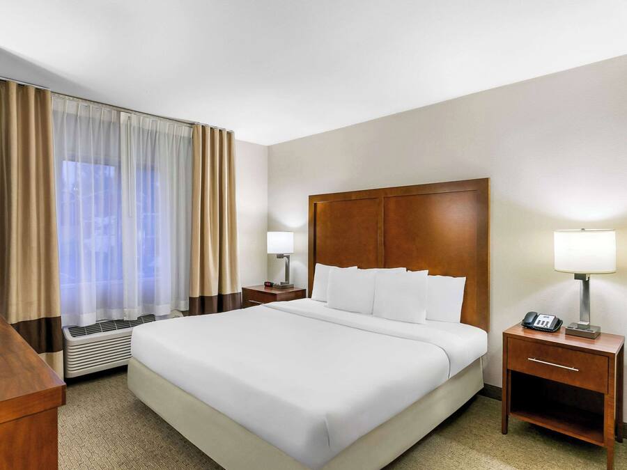 A hotel room with a large bed, a wooden nightstand with a lamp, a heater, and a window with curtains.