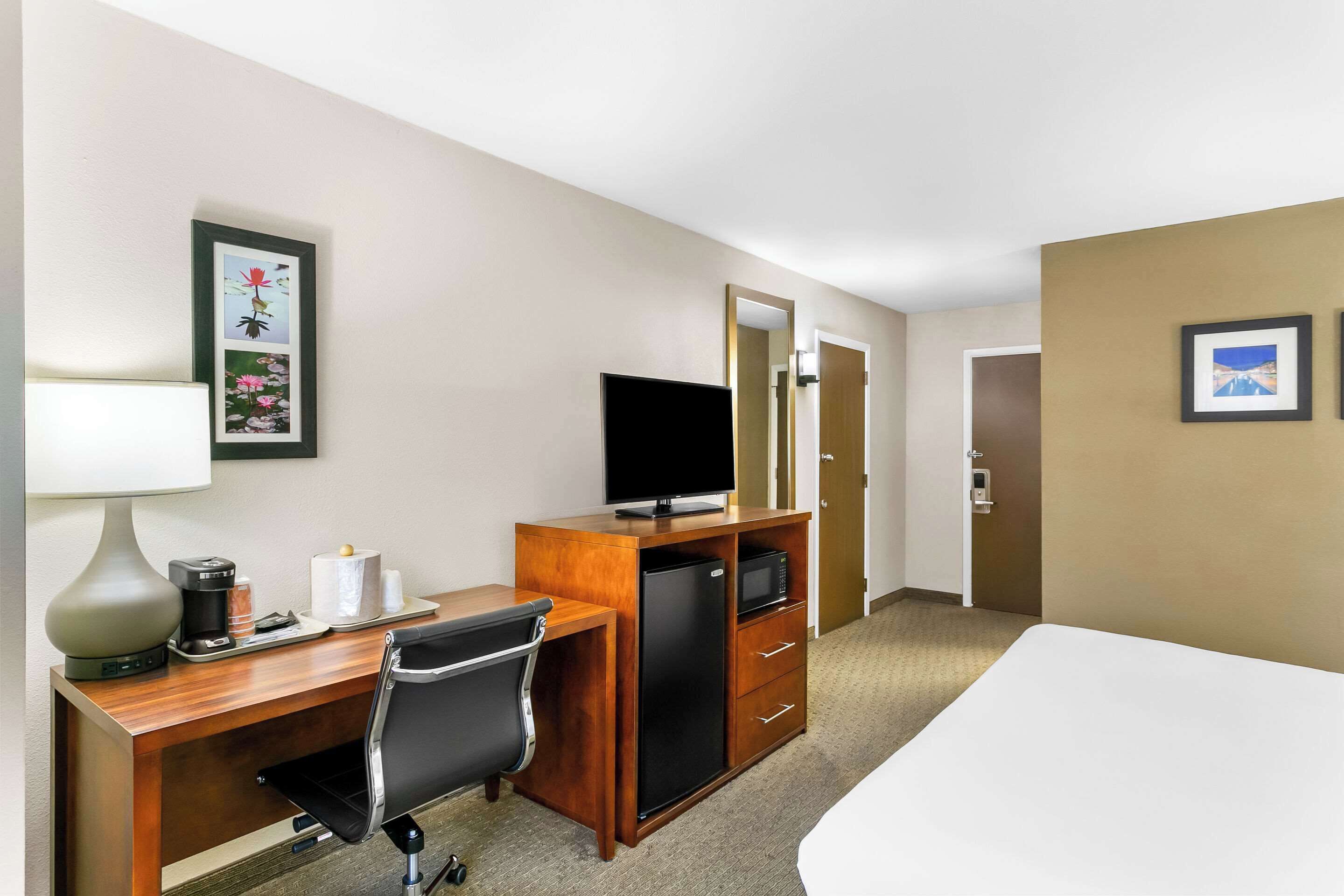 A hotel room with a bed, a desk with a computer, a refrigerator, a microwave, a lamp, and a chair.