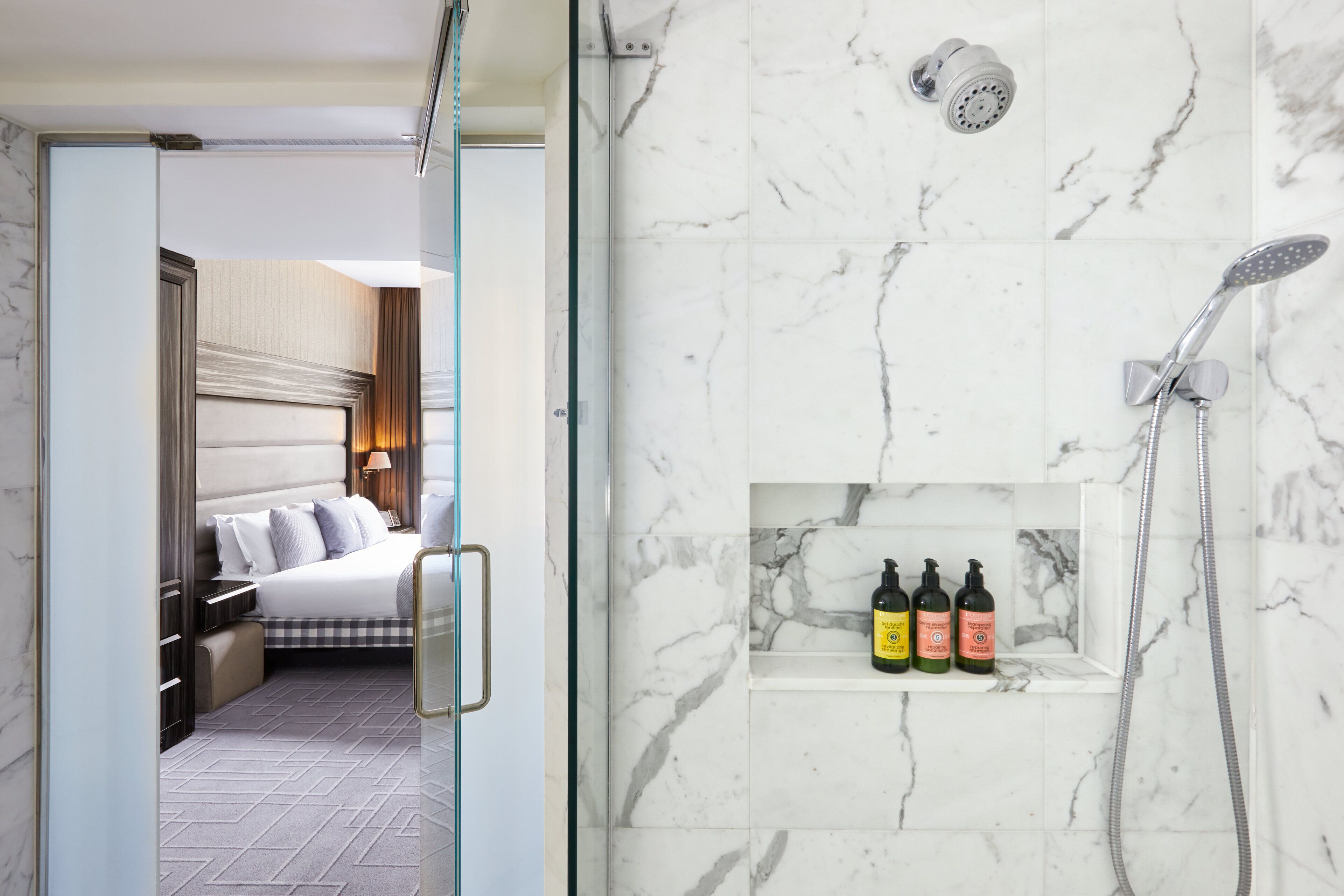 A hotel room with a bed, a bedside table, a nightstand, and a bathroom with a shower, a shelf with toiletries, and a marble wall.