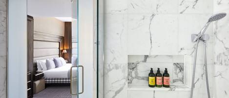 A hotel room with a bed, a bedside table, a nightstand, and a bathroom with a shower, a shelf with toiletries, and a marble wall.