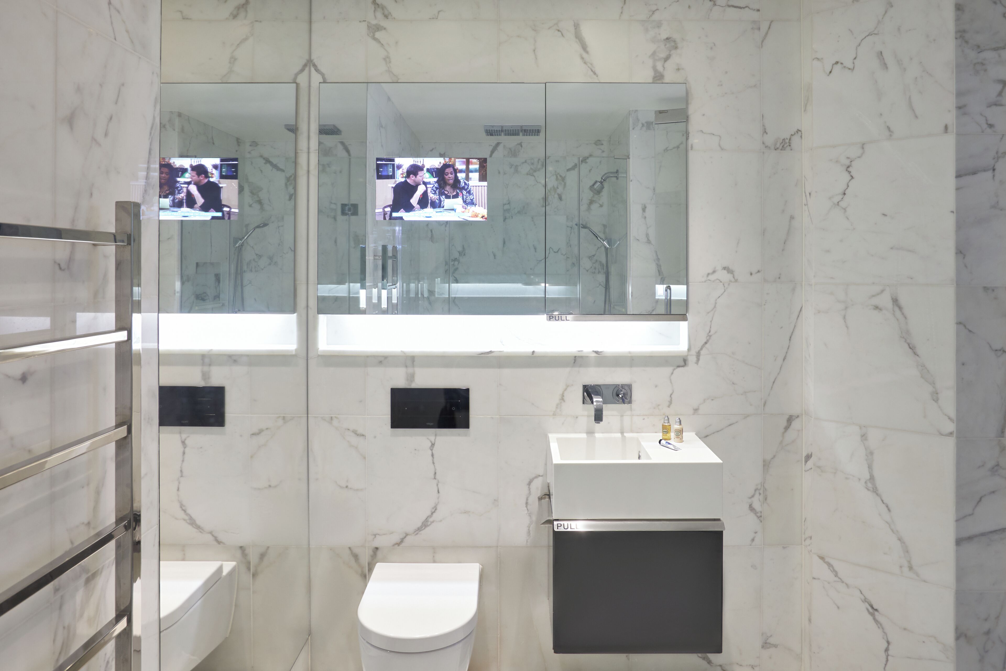 A modern bathroom with a glass partition, a white toilet, a sink with a mirror, and a television screen.