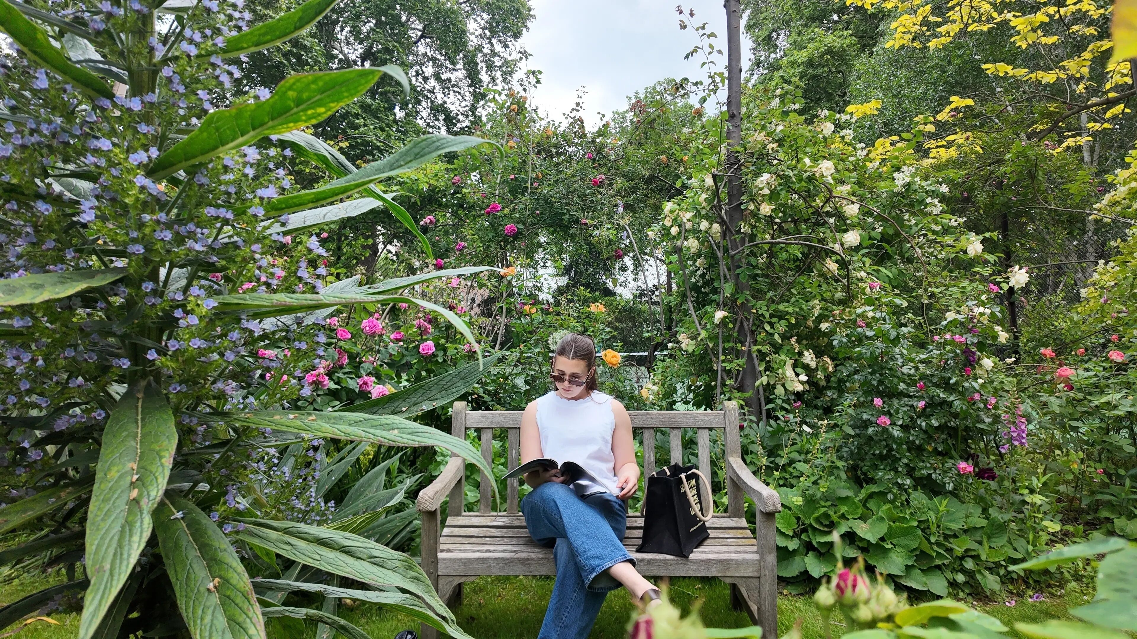Une personne assise sur un banc en bois, entourée d’une végétation luxuriante et de plantes en fleurs.