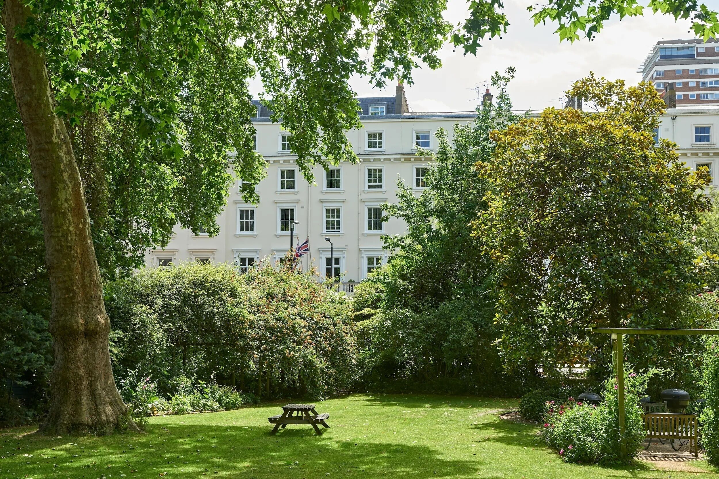 A lush garden with a picnic table, dense foliage, and a building with multiple windows in the background.