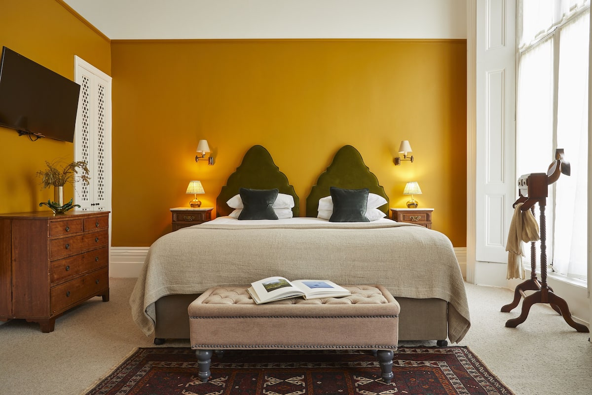 A bedroom with a large bed, two nightstands with lamps, a dresser, a bench with a book, and a window with curtains.