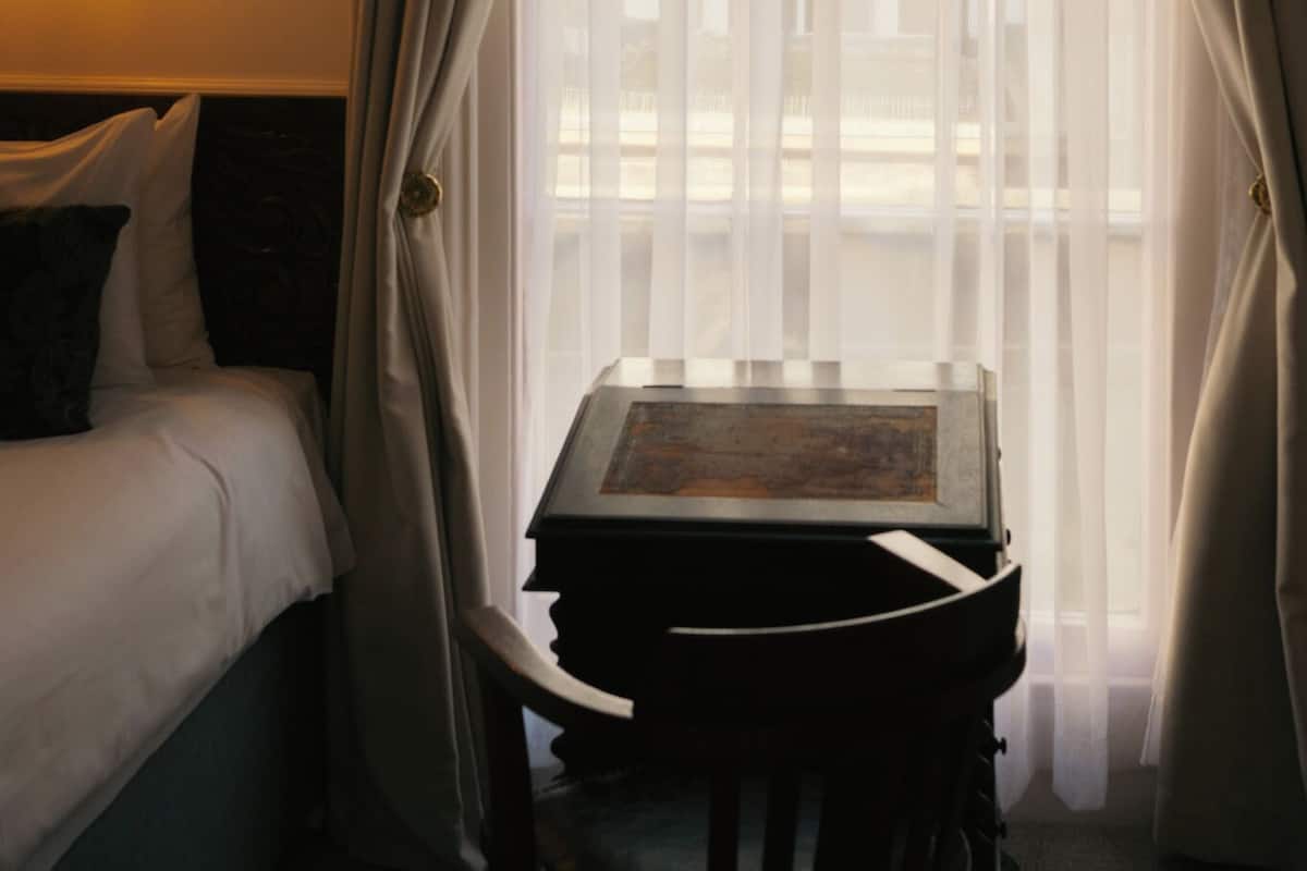 A hotel room with a bed, a table with a chair, and sheer curtains.