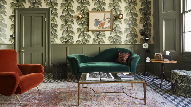 A room with a green velvet sofa, a red armchair, a glass coffee table with books, a patterned rug, and a wall with a tree wallpaper. - Georgian House Hotel (London)