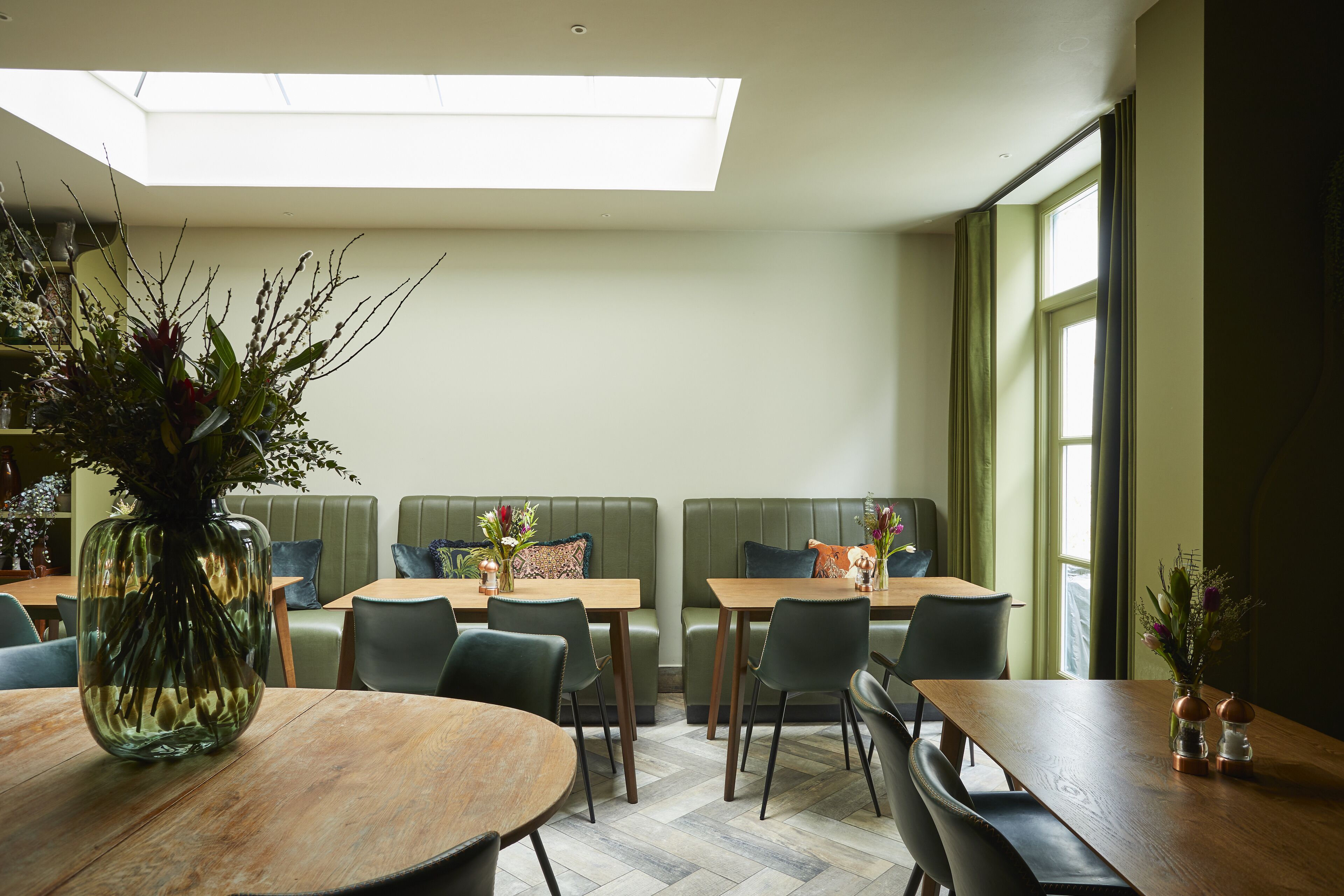 A restaurant with a high ceiling and skylight, green upholstered booths, wooden tables, and chairs.