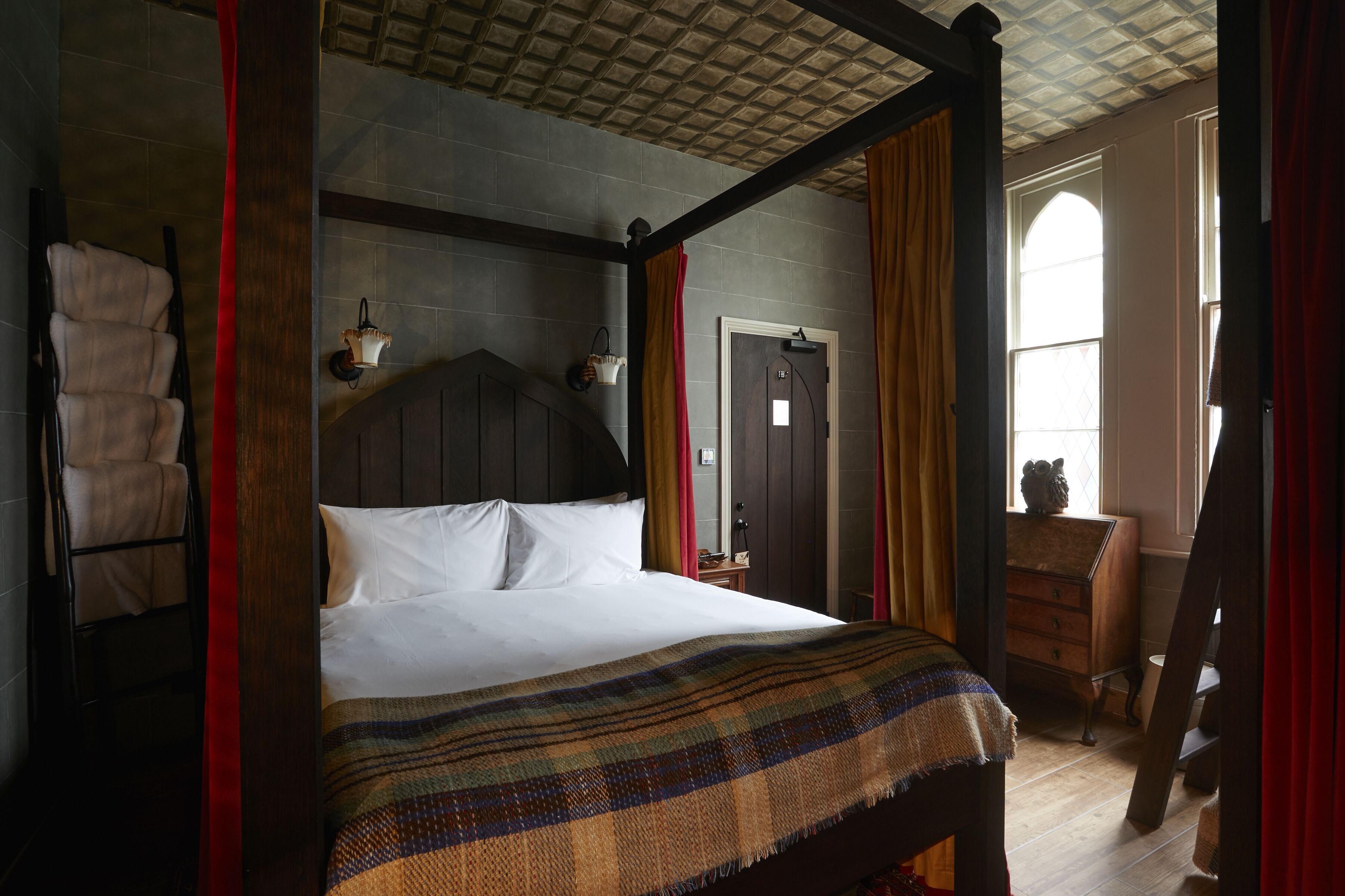 A four-poster bed with a plaid blanket, a wooden dresser, and a ladder in a room with a checkered ceiling and a window with curtains.