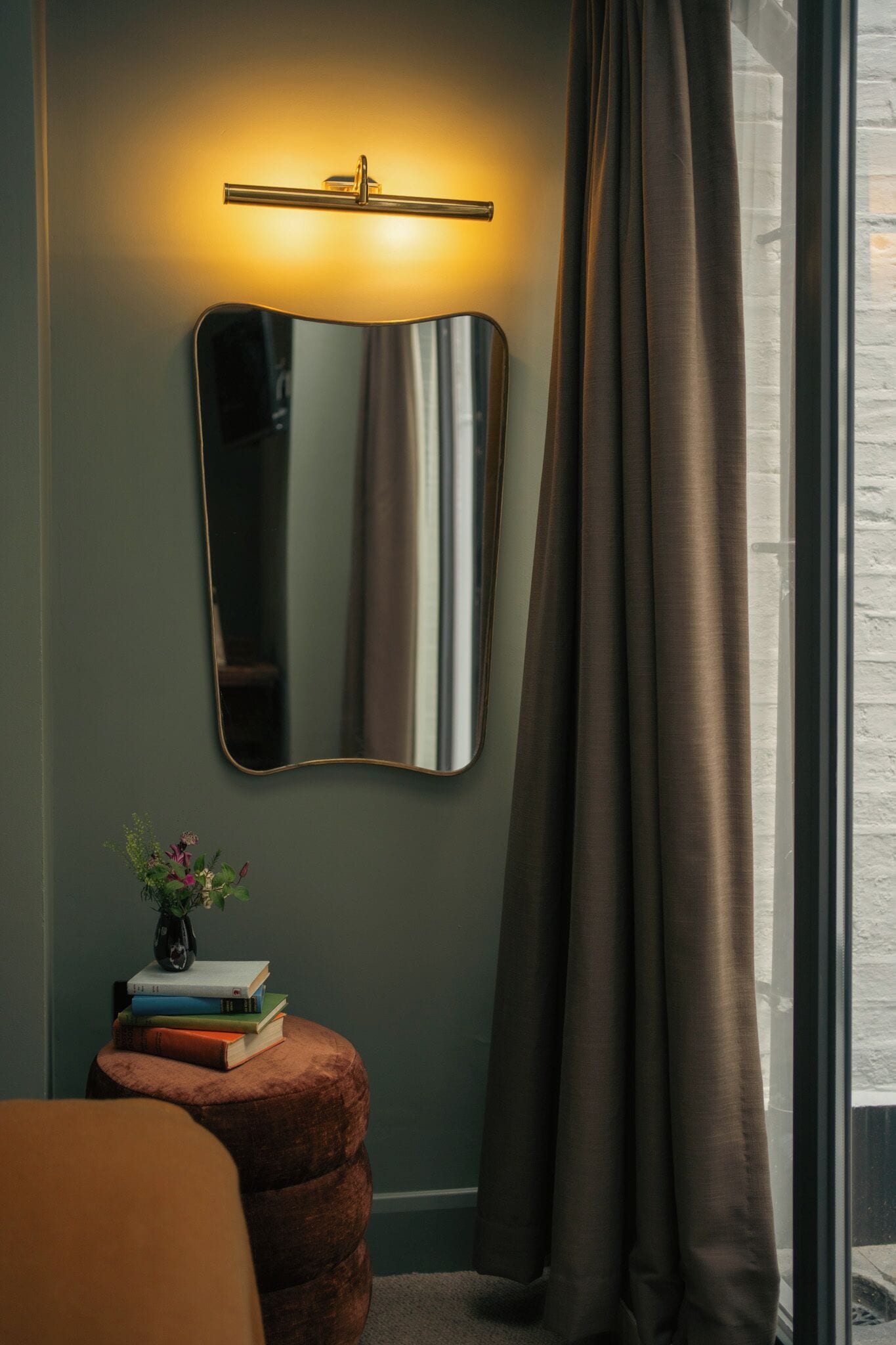 A room with a wall-mounted mirror, a bedside table with books, a small plant, and a window with curtains.