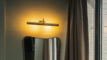 Ein Zimmer mit einem Wandspiegel, einem Nachttisch mit Büchern, einer kleinen Pflanze und einem Fenster mit Vorhängen.