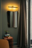 A room with a wall-mounted mirror, a bedside table with books, a small plant, and a window with curtains.