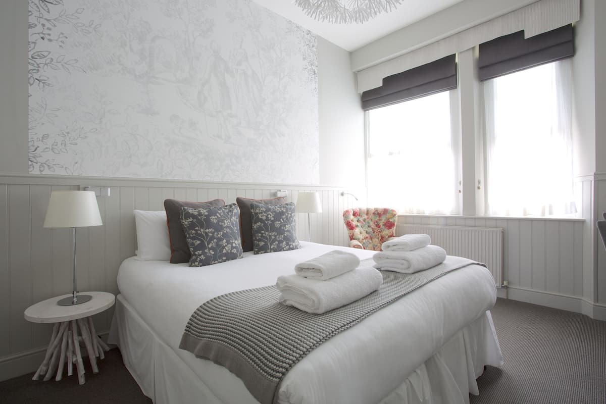 A neatly arranged hotel room with a large bed, two bedside lamps, a round side table, a floral armchair, and a view of the outdoors through the window.