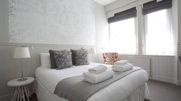 Habitación de hotel impecable con una cama grande, dos lámparas de noche, una mesita redonda, un sillón con estampado floral y vistas al exterior a través de la ventana.