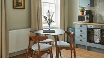 Een eetgedeelte met een ronde tafel, twee stoelen, een vaas met bloemen, een raam met gordijnen, en een keuken met kasten en apparatuur.