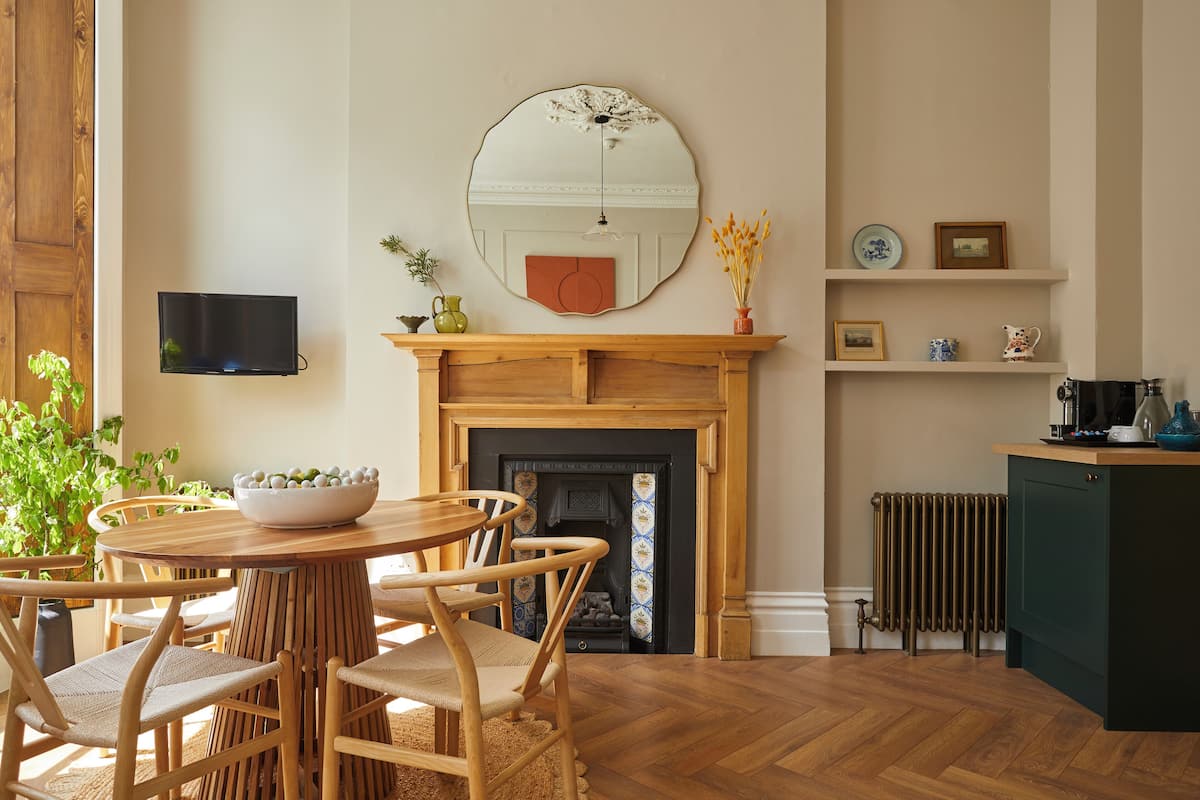 A dining area with a round wooden table, chairs, and a bowl of fruit. There is a fireplace with a mantel, a mirror, and a television mounted on the wall.