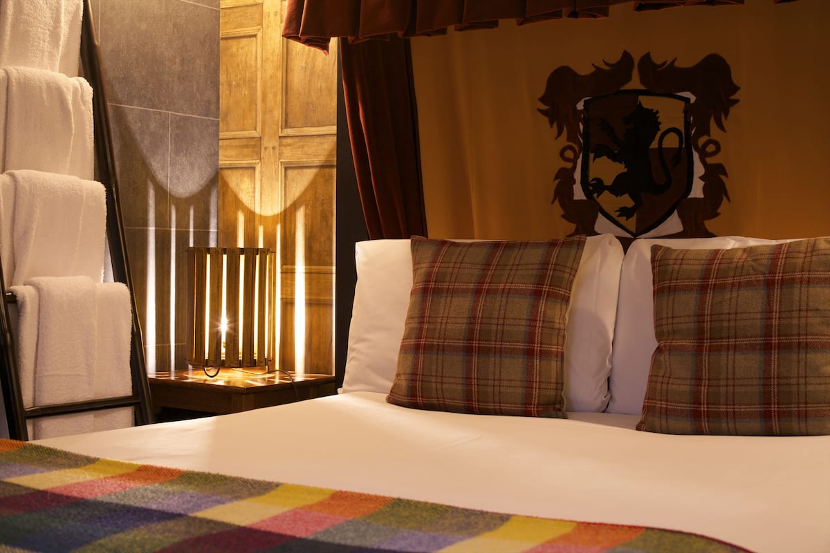 A hotel room with a bed, a nightstand, a lamp, a towel rack, and a wall with a coat of arms.