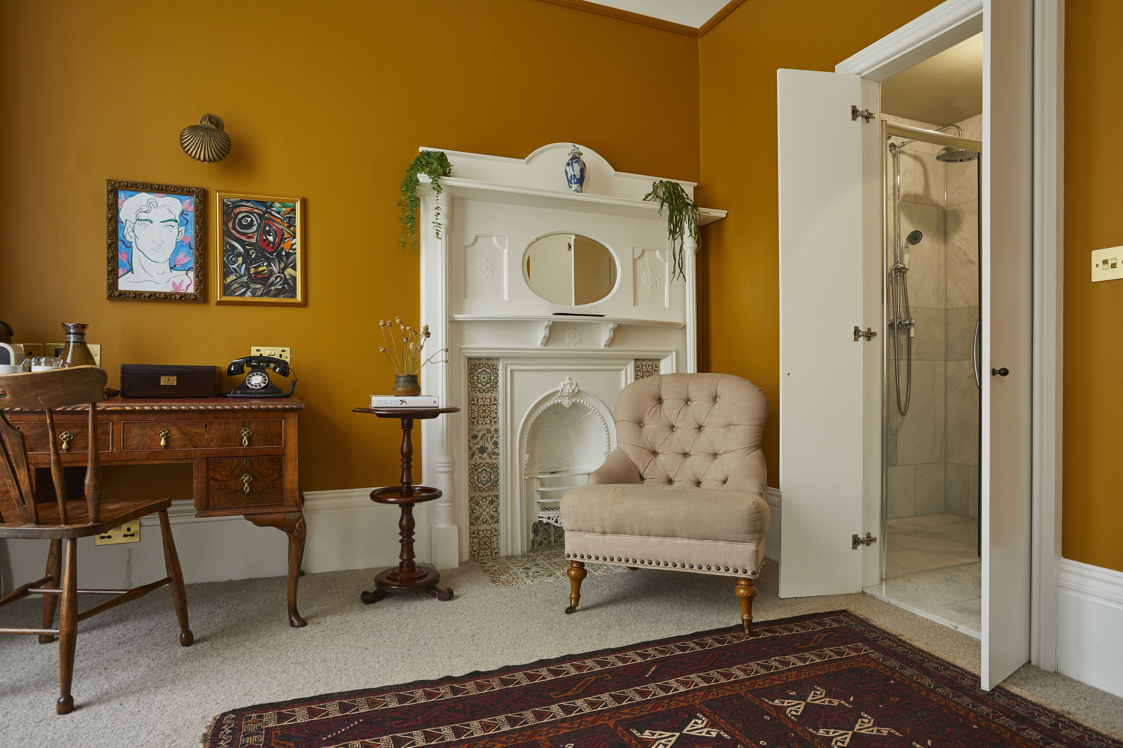 A room with a wooden desk, a beige armchair, a fireplace, and a shower area.