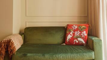 Un sofá verde con un cojín rojo, una mesa de madera con un cuenco de manzanas verdes y un libro.