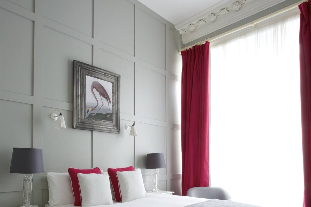 A hotel room with a large bed, two bedside tables, a nightstand, a chair, and a window with red curtains.