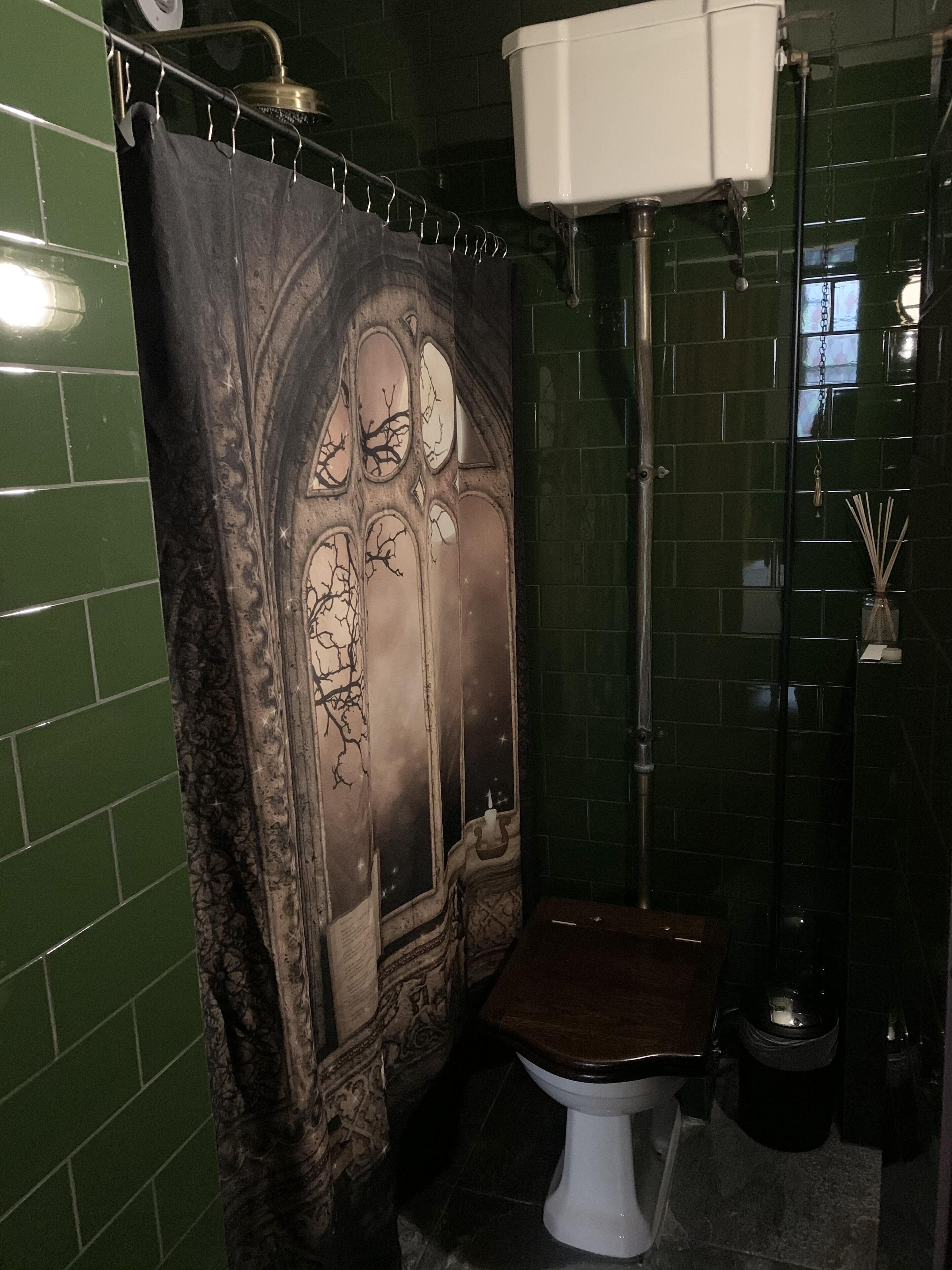 Un baño con cortina de ducha de temática gótica, un inodoro blanco y paredes con azulejos verdes.