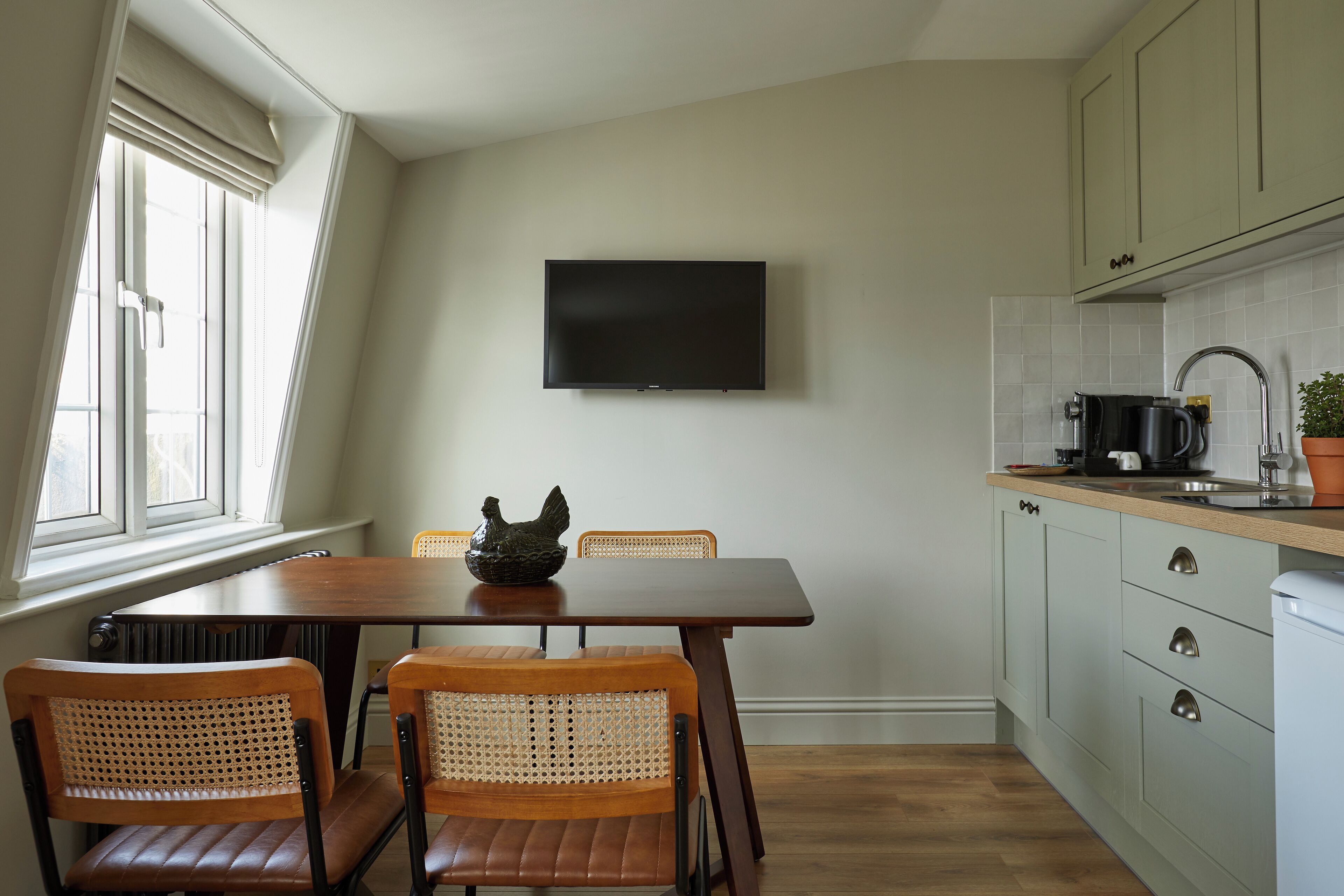 Een keuken met een eettafel, stoelen, een flatscreen televisie, kasten en een raam.