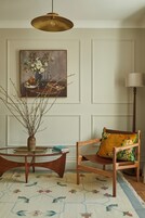 Et værelse med et rundt træbord, en stol med blomstret pude, en vase med grene, et billede på væggen og en gulvlampe.