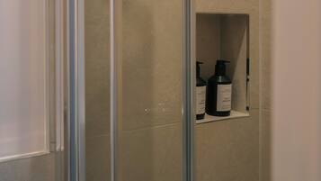 Un baño con cabina de ducha de vidrio, un estante con artículos de aseo y un muro con azulejos.