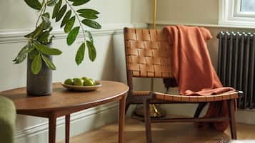 Una silla de madera con respaldo tejido y una manta sobre ella, una mesa de madera redonda con un bol de manzanas verdes, y una planta en maceta con hojas anchas.
