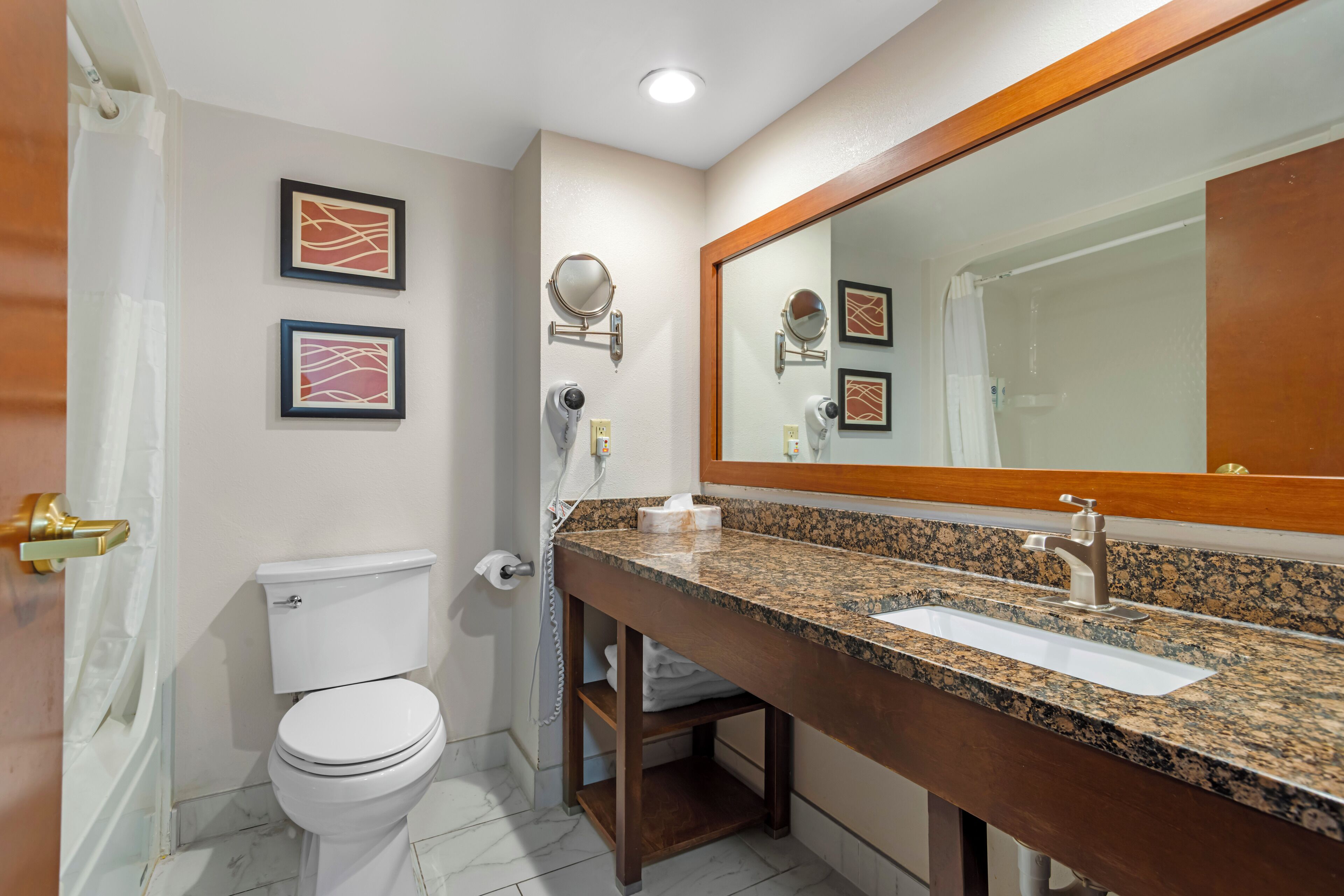 A bathroom with a white toilet, a sink with a granite countertop, a mirror with a wooden frame, and two framed pictures on the wall.
