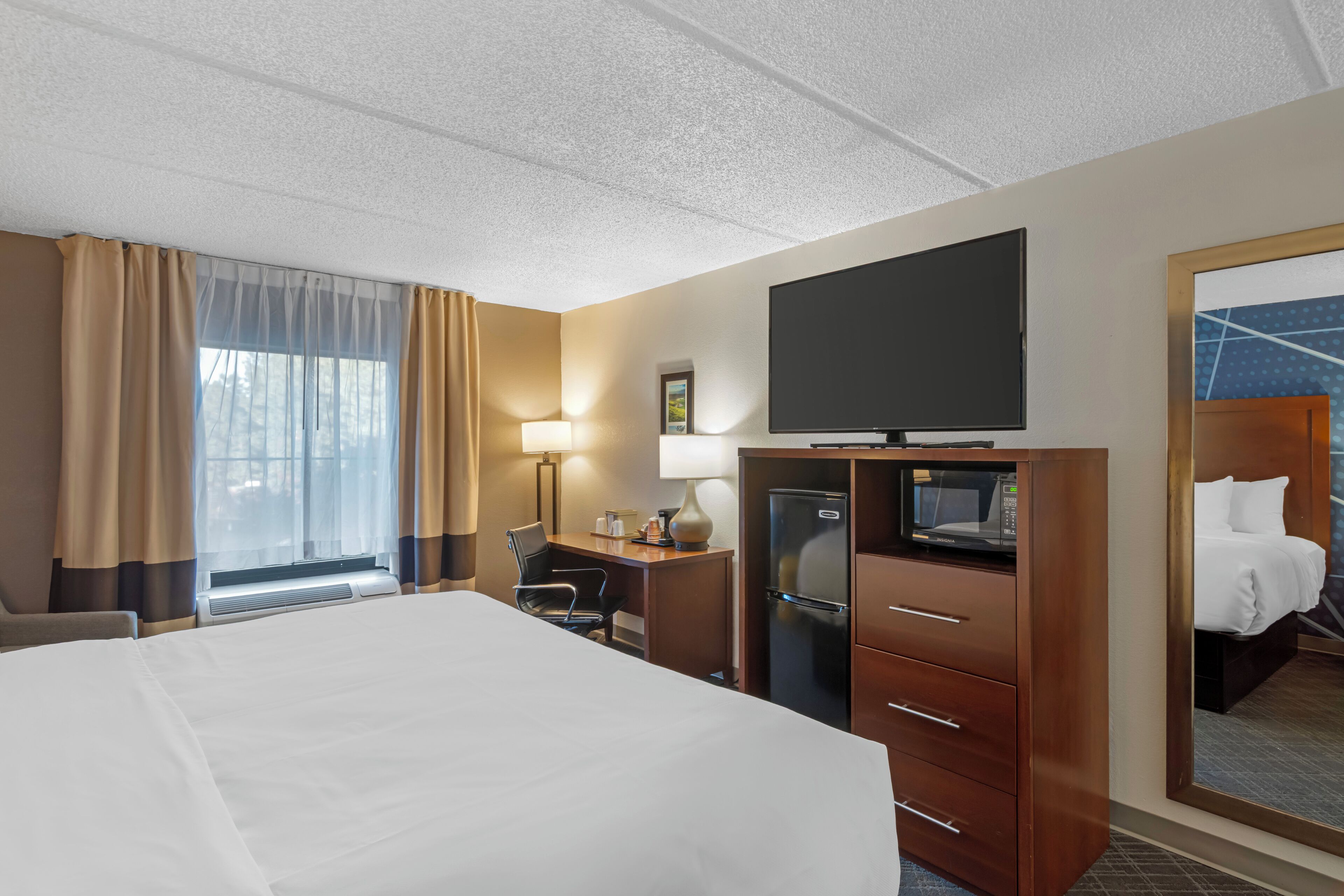 A hotel room with a large bed, a desk with a chair, a flat-screen TV, a refrigerator, and a mirror reflecting the bed.
